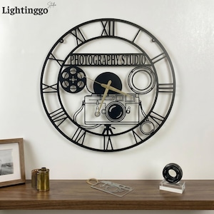 May include: A circular black metal wall clock featuring Roman numerals and a central design of a camera, a film reel, and a lens. The text "PHOTOGRAPHY STUDIO" is cut out in the upper section. The clock has thin gold-colored hands.