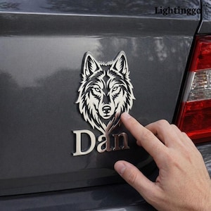 Peut inclure: Un emblème argenté en forme de tête de loup avec le nom "Dan" en dessous, fixé sur une voiture grise. Le design du loup est détaillé, avec un accent sur les yeux et la fourrure. Un doigt touche l'emblème.