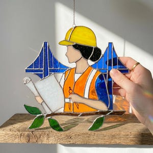 Peut inclure: Vitrail représentant une femme ingénieur en bâtiment portant un casque jaune et un gilet orange, tenant des plans. Le design comprend un pont bleu et des feuilles vertes, suspendu à une chaîne. L'œuvre est sur fond neutre.