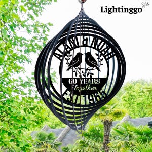 May include: Black metal wind spinner with a spiral design. The center features the names "ALAN & NOAH" above two birds and the text "60 YEARS Together EST. 1963". The spinner hangs outdoors with a green foliage background.