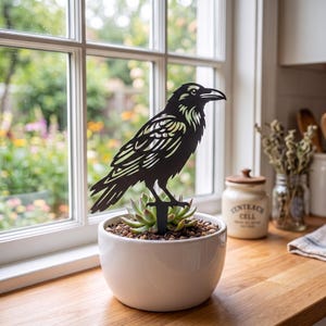 May include: A black metal raven silhouette garden stake is inserted into a white ceramic planter containing a green succulent plant. The raven figure features intricate cut-out details on its wings and feathers, standing upright on a thin metal stem.