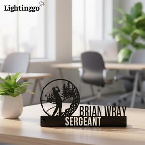 Pode incluir: Placa de mesa de metal preto com um design temático de golfe. Apresenta uma silhueta de golfista, árvores e o texto "BRIAN WRAY SERGEANT". A placa é exibida em uma mesa de madeira.