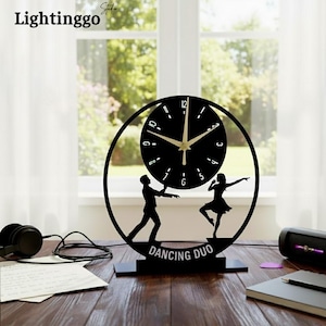 Könnte beinhalten: Schwarze dekorative Uhr mit kreisförmigem Design, das ein Zifferblatt und Silhouetten eines tanzenden Paares zeigt. Die Uhr hat goldfarbene Zeiger und den Text "DANCING DUO" an der Basis. Der Markenname "Lightinggo" befindet sich oben links.