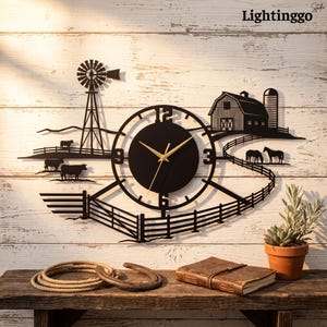 May include: A black metal wall clock featuring a farm scene with a windmill, a barn, a silo, and cattle. The clock face is circular with gold-coloured hands and large numerals. A fence design extends across the bottom, connecting the elements.
