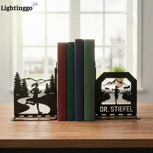 May include: Black metal bookends with a running theme. One depicts a runner on a path through a forest, the other a runner crossing a finish line. The bookends are supporting a row of books.