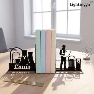 Op de afbeelding: Een zwarte metalen boekensteunset met het silhouet van een kerncentrale met de naam Louis aan de ene kant en een bouwvakker met een blauwdruk aan de andere kant, ondersteunend vier pastelkleurige boeken op een houten oppervlak.
