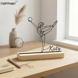 May include: A black metal gymnast figure holding a ball, mounted on a wooden base. The name "Katy" is written in cursive on the base. The figure is in a graceful pose, with one leg extended. A vase with dried flowers is in the background.