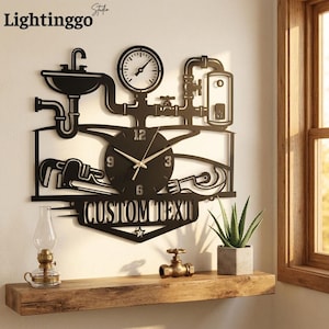 Pode incluir: Um relógio de parede de metal preto com um design de canalização, incluindo um lavatório, tubos, um manómetro e uma chave inglesa. O mostrador tem ponteiros dourados e os números 3, 6, 9 e 12. Abaixo, uma faixa exibe o texto CUSTOM TEXT.
