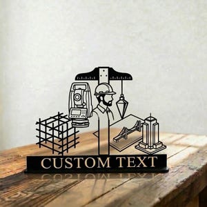 May include: Black metal desk sign featuring an engineer design with a total station, bridge, and skyscraper illustrations. The sign includes the text "CUSTOM TEXT" on a rectangular base, suitable for personalization.