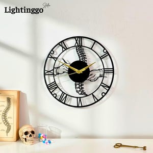 May include: Black metal wall clock with Roman numerals and gold hands. The clock features a spine design with hands and a black center. The clock is mounted on a white wall.