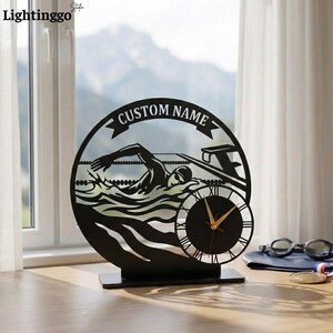 Könnte beinhalten: Eine schwarze Metall-Schreibtischuhr mit Schwimmer-Design. Das Zifferblatt hat römische Ziffern und goldene Zeiger. Die Worte "CUSTOM NAME" stehen über dem Schwimmer. Die Uhr steht auf einem schwarzen Sockel.