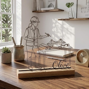 Puede incluir: Una escultura de alambre negro sobre una base de madera rectangular que representa a un trabajador de la construcción con casco y chaleco, de pie ante un escritorio con planos, un modelo de puente y un nivel. El nombre Olive está en cursiva.