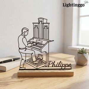 Puede incluir: Una escultura de alambre negro que representa a una persona tocando un órgano de tubos, montada sobre una base de madera rectangular. La escultura integra el nombre Philippe en letra cursiva en el diseño, mostrando una escena musical.