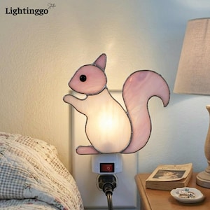 Puede incluir: Una luz de noche de ardilla rosa y blanca enchufada a una toma de corriente. La lámpara con forma de ardilla tiene un diseño de vidrieras con una luz que brilla a través del cuerpo. La lámpara está al lado de una cama con un edredón estampado.