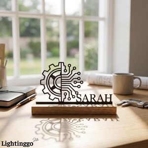 May include: A black laser-cut desk nameplate with the name "SARAH". The design features a gear and circuit board symbol. The nameplate sits on a wooden base. A mug, notebook, and other office supplies are in the background.