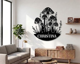 May include: Black metal wall art depicting a group of mushrooms with dotted caps and the name "CHRISTINA" in white. The artwork is mounted on a white wall, with a living room setting in the background.