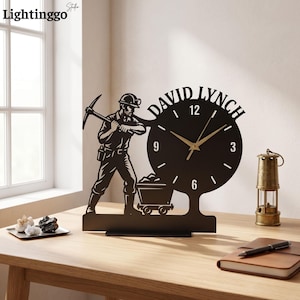 May include: A black metal desk clock featuring a silhouette of a miner holding a pickaxe next to a mine cart. The clock face displays the name DAVID LYNCH in bold letters at the top, with gold-coloured hour and minute hands and white markers.