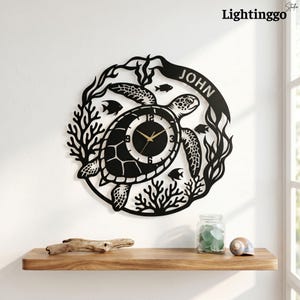 May include: A circular black metal wall clock featuring a sea turtle design surrounded by coral and small fish. The clock face is integrated into the turtle shell and includes the name JOHN on a curved banner at the top with gold-colored hands.