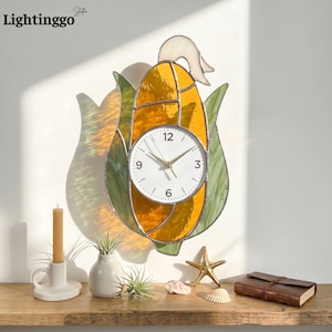 May include: A stained glass wall clock shaped like an ear of corn, featuring a white clock face with black numerals and gold hands. The clock is yellow and orange with green leaves. The word "Lightinggo" is in the top left corner.