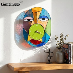 Puede incluir: Un reloj de pared ovalado con una esfera de vidrieras con un patrón geométrico multicolor que forma un rostro estilizado. El centro tiene una esfera de color verde lima con manecillas doradas y números blancos en las posiciones 12, 3, 6 y 9.