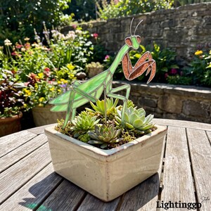 May include: A stained glass praying mantis sculpture in shades of light green and copper, mounted on a thin metal rod. The sculpture is positioned in a square, off-white ceramic planter filled with several small green succulent plants.