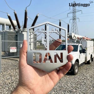 以下が含まれることがあります： 電線作業員が電柱を登る様子を描いたシルバーの金属製切り抜き。下部には「DAN」という名前が切り抜かれています。背景には送電線、ユーティリティトラック、そして「Lightinggo」の文字があります。