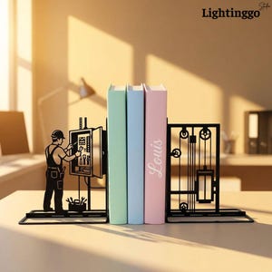 Op de afbeelding: Een paar zwarte metalen boekensteunen met het silhouet van een elektricien die aan een open schakelkast werkt aan de ene kant en een mechanisch katrolsysteem aan de andere kant, die drie boeken met pastelkleurige kaften vasthouden.