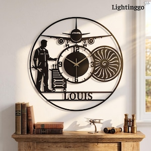 May include: A circular black metal wall clock featuring a silhouette of an airplane, a jet engine, and a mechanic holding a wrench next to a tool cart. The clock face has numbers 3, 6, 9, and 12, with the name LOUIS displayed in capital letters.