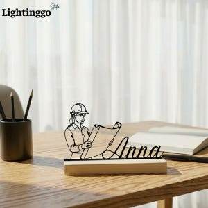 Peut inclure: Un porte-nom de bureau en bois avec une silhouette noire d'une architecte portant un casque de chantier, tenant un plan. Le nom "Anna" est écrit en cursive. Le porte-nom est sur une base en bois.