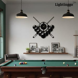 Könnte beinhalten: Eine schwarze Wanduhr mit Billard-Motiv, mit einem Zifferblatt, das von Billardkugeln und gekreuzten Queues umgeben ist. Das Wort "Lightinggo" befindet sich in der oberen rechten Ecke. Die Uhr hängt über einem Billardtisch.