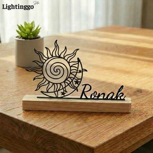 May include: A black metal sun and moon desk decoration with the name "Ronak" on a wooden base. The sun has a spiral center and the moon is adorned with stars. A small potted succulent is in the background.