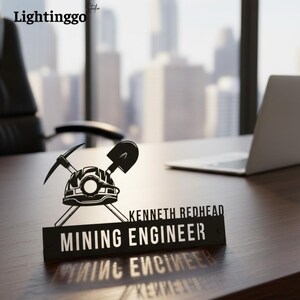 May include: A black desk nameplate with the text "MINING ENGINEER" and the name "KENNETH REDHEAD". The nameplate features a mining helmet, pickaxe, and shovel design. The nameplate is on a wooden desk.