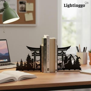 Könnte beinhalten: Ein Paar Buchstützen aus schwarzem Metall mit einer lasergeschnittenen Silhouette eines traditionellen Torii-Tors, Kiefern und Berggipfeln. Die Buchstützen stehen auf einem Schreibtisch aus Holz und stützen drei aufrecht stehende Bücher.