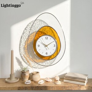 May include: A modern wall clock with a white face, gold hands, and black numerals. The clock is set within a yellow, egg-shaped frame, surrounded by a silver metal outline. The word "Lightinggo" is in the top left corner.