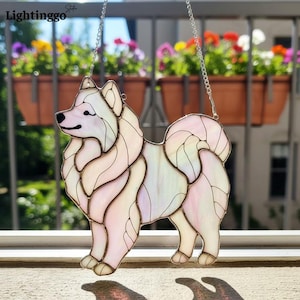 May include: A stained glass dog suncatcher in the shape of a Samoyed. The dog is primarily white with pastel pink and yellow accents. It hangs from a silver chain. The word "Lightinggo" is in the top left corner.