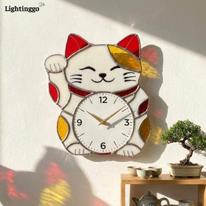 May include: A white, red, and yellow Maneki-neko cat-shaped wall clock. The clock face is white with black numbers and gold hands. The cat is smiling and has one paw raised. The clock is hanging on a white wall.