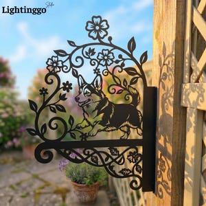 Könnte beinhalten: Schwarzes Metallschild mit einem Corgi-Hund-Design, umgeben von floralen und Ranken-Akzenten. Das Schild ist an einem Holzpfosten befestigt. Das Wort "Lightinggo" ist in der oberen linken Ecke sichtbar.