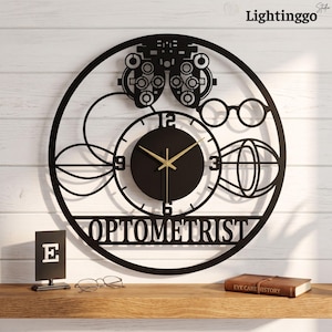 May include: A circular black wall clock featuring a central dial with gold hands and the word OPTOMETRIST in bold lettering. The design includes cutouts of an ophthalmic phoropter, eyeglasses, and abstract eye-related geometric shapes.