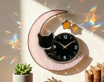 May include: A decorative wall clock featuring a black cat silhouette resting on a pink crescent moon. The clock has a black face with gold hands and a gold star charm. The clock is approximately 25 cm in diameter.