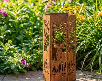 May include: A weathered, hexagonal metal lantern with a deer and forest silhouette design. The lantern is made of brown metal and has cut-out details. The background features lush greenery and flowers. The text "Lightinggo" is in the top left corner.