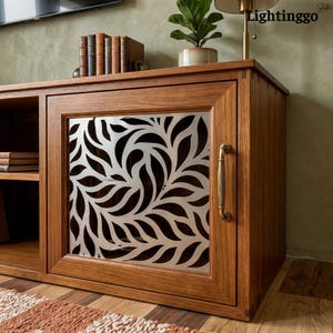 May include: A wooden media console featuring a cabinet door with a silver-toned metal panel displaying a laser-cut leaf pattern. The cabinet includes a vertical brass-coloured handle, exposed joinery details, and an open shelf with stacked books.