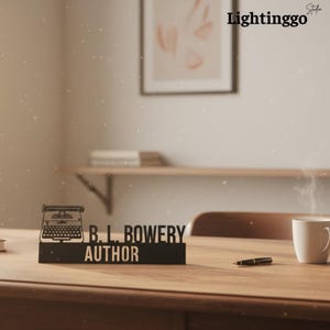May include: A black desk sign with the text "B.L. BOWERY AUTHOR" and a typewriter silhouette. The sign is on a wooden desk with a pen and a white mug. The image also includes the text "Lightinggo" in the upper right corner.