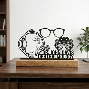 Op de afbeelding: Een zwart, lasergesneden houten bureau-ornament voor een oogarts. Het ontwerp omvat een oog, een bril, een phoropter en de tekst "DR. JOHN DAVIS OPHTHALMOLOGY" op een houten voet.