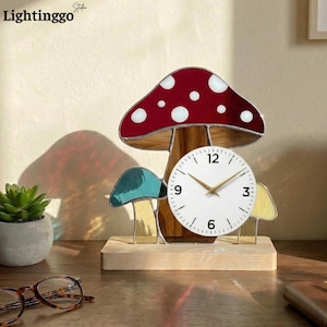 Puede incluir: Un reloj decorativo con forma de hongo con una tapa roja con lunares blancos, detalles turquesa y amarillos y una esfera blanca. El reloj está montado sobre una base de madera. La marca "Lightinggo" está en la esquina superior izquierda.