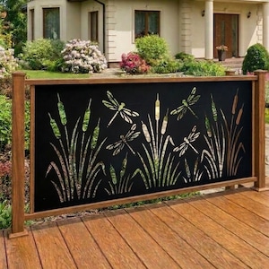 May include: A black metal decorative railing panel featuring a laser-cut design of dragonflies and cattails. The rectangular panel is framed by wooden posts and rails, positioned on a wooden deck with a house and garden visible in the background.
