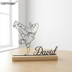 Könnte beinhalten: Schwarze Metallskulptur eines Breakdancers im Handstand, montiert auf einem Holzsockel. Der Name "David" ist in Schreibschrift geschrieben. Die Skulptur ist ein dekorativer Artikel.