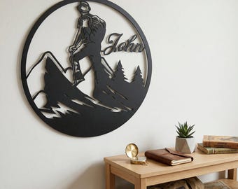 May include: Black metal wall art showing a hiker climbing a mountain, framed in a circle. The name "John" is written across the design. The art piece features mountain and tree silhouettes, ideal for home decoration.