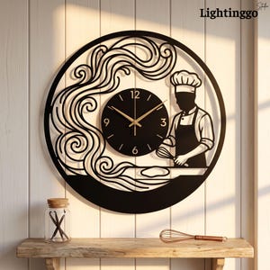 May include: A circular black metal wall clock featuring a silhouette of a chef wearing a toque and apron, holding a whisk over a counter. Swirling decorative lines extend from the chef. The clock face has gold hands and numerals 3, 6, 9, 12.
