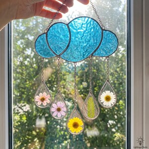 May include: A stained glass cloud with five teardrop-shaped pendants. Each pendant contains a pressed flower or leaf. The cloud is a textured, light blue color, and the pendants hang from silver chains. A unique floral decoration.