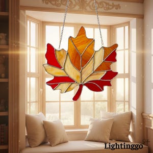 May include: A stained glass maple leaf decoration with red, orange, and yellow hues, suspended by a silver chain. The leaf is composed of multiple glass pieces, creating a mosaic effect. The background shows a window with a cozy seating area.
